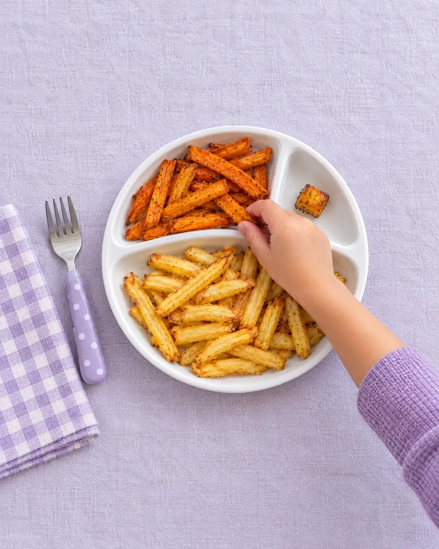 Prato com batata frita, batata-doce em palito e um pedacinho de abóbora, com a mão de uma criança tocando a batata-doce